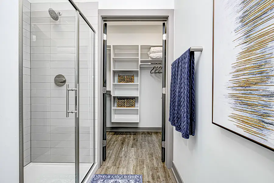 A bathroom with a shower on the left and towel rack and abstract artwork on the right. Then facing an open walk in closet with shelving and hangers